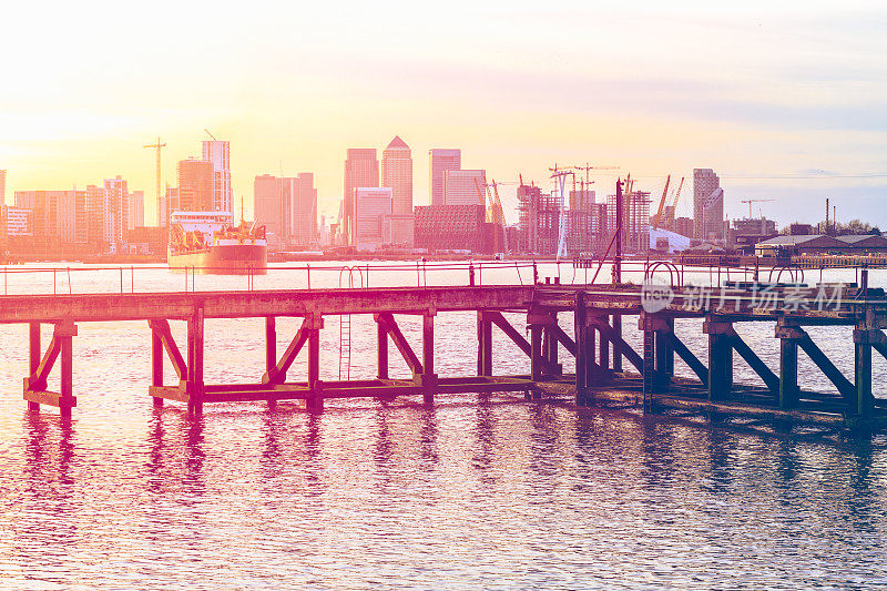 夕阳在伦敦一个废弃的码头上投下粉红色的光，背景是一艘靠近的船和金丝雀码头