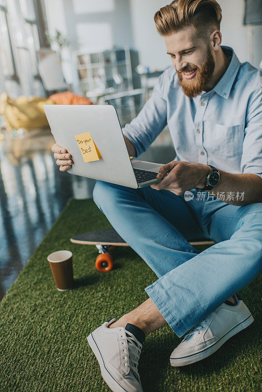 微笑的大胡子男人坐在滑板上，使用笔记本电脑