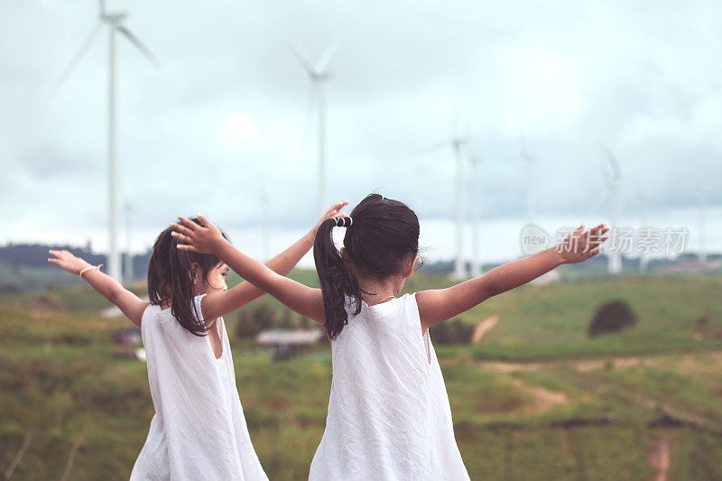 后视图的两个亚洲儿童女孩举起他们的手臂看风力涡轮机场