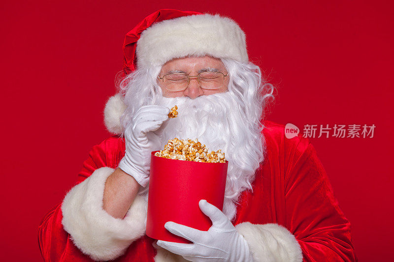 传统的圣诞老人看电视，吃爆米花。圣诞节。红色的背景。情感恐惧惊喜