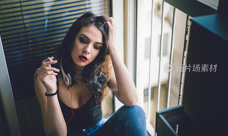一个戴着耳机的女人坐在窗边抽烟。
