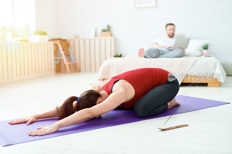 年轻女子在运动服装躺在孩子的姿势和做补偿运动，同时练习瑜伽在家，而丈夫在床上工作