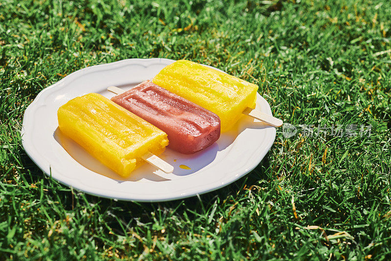 三根冰棍放在一个白色的盘子里，在一个炎热的阳光灿烂的夏天的花园草坪上。
