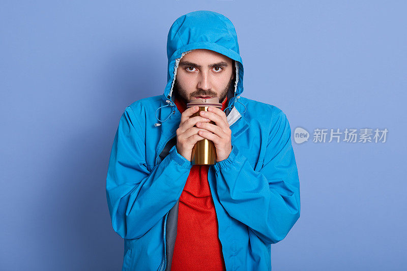 摄影棚拍摄的有吸引力的年轻男子有胡子，有不开心的面部表情，拿着热杯和咖啡或茶，穿着蓝色夹克和红衫，冷，晒手。