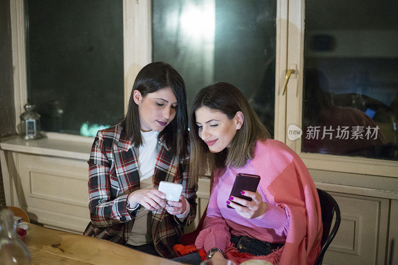 两个女人在咖啡馆里玩得很开心，还在看智能手机。一位女士向她坐着的朋友展示某样东西。