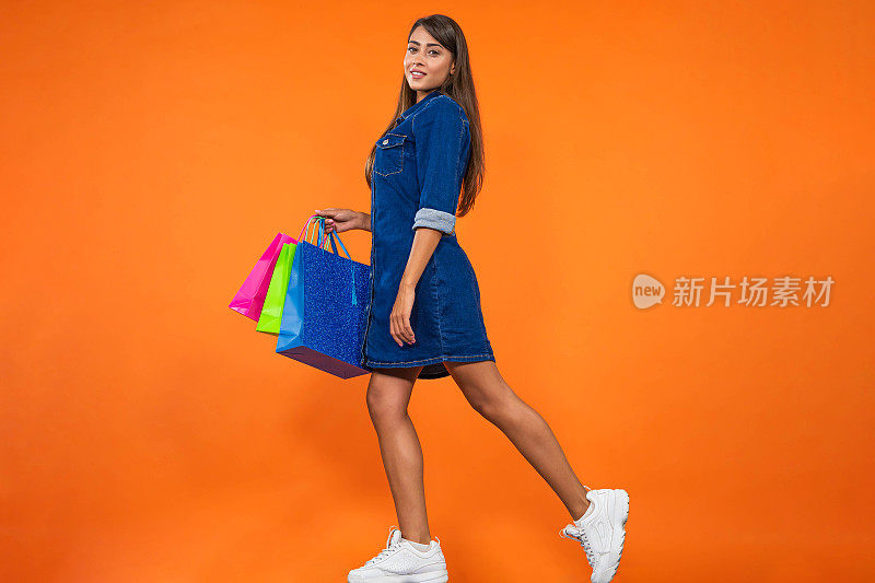 年轻美丽的女人在牛仔裙携带购物袋在橙色的背景。购物概念，夏季大甩卖。