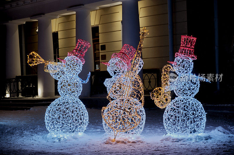 三个由金属丝和花环组成的彩色发光的雪人在冬夜的城市街道上演奏爵士乐。