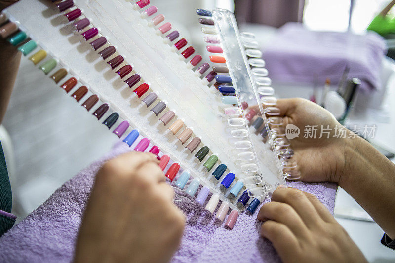 年轻女子看指甲颜色调色板-彩色的人工指甲在一个美甲店-一个年轻女子选择她的指甲的颜色，而美容师帮助她选择-指向指甲油的颜色