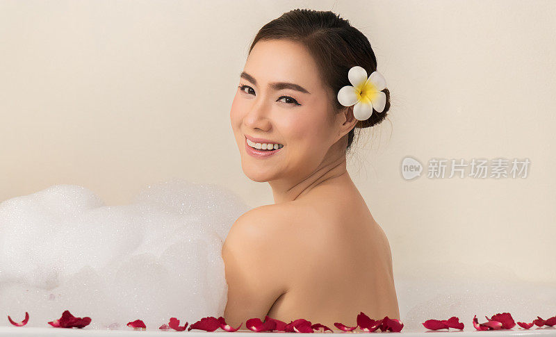 放松美丽的亚洲女人纵容她的身体在温泉浴缸与白色泡沫泡沫的肥皂和玫瑰花瓣。休闲的追求