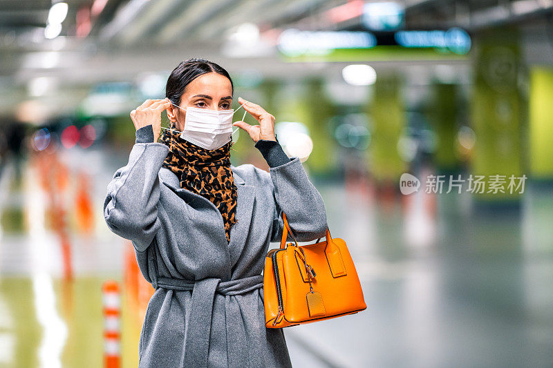 在新冠肺炎大流行期间，身着灰色外套、橙色手袋、印花围巾的女子在停车场戴上防护口罩