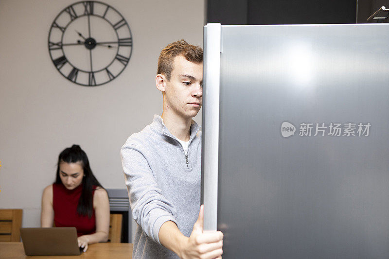 一名男大学生正在打开冰箱门，一名女大学生正在厨房桌子上用笔记本电脑工作