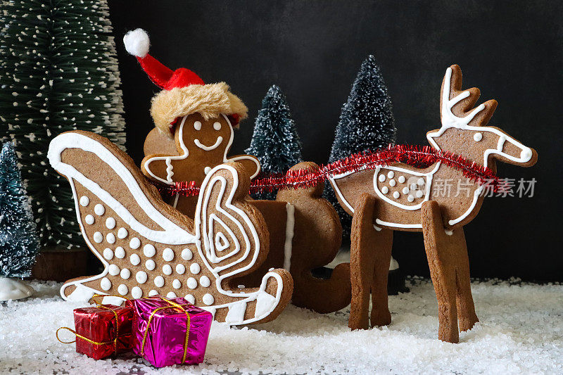 近景自制，姜饼饼干圣诞老人，驯鹿和雪橇，装饰着白色的皇家糖衣，圣诞老人帽，圣诞礼物包围的模型杉和雪的夜间圣诞森林场景，黑色的背景