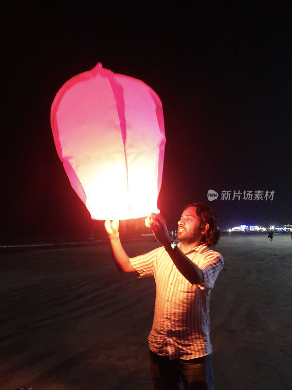 印度男子举着点燃的中国纸灯笼，在海边放纸灯笼的特写，聚焦于前景