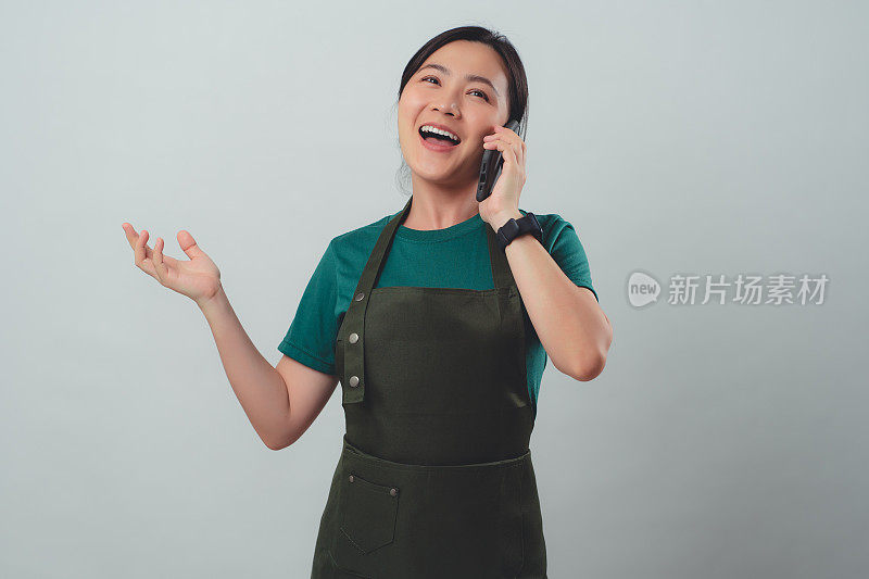 一名穿着围裙的亚洲女子开心地微笑着，用智能手机打电话给客户确认订单。