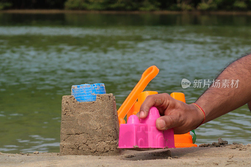 特写照片，一个无法辨认的人在泻湖水边的沙滩上用塑料堆沙堡，色彩鲜艳的儿童沙滩玩具，水桶和铁锹，重点放在前景