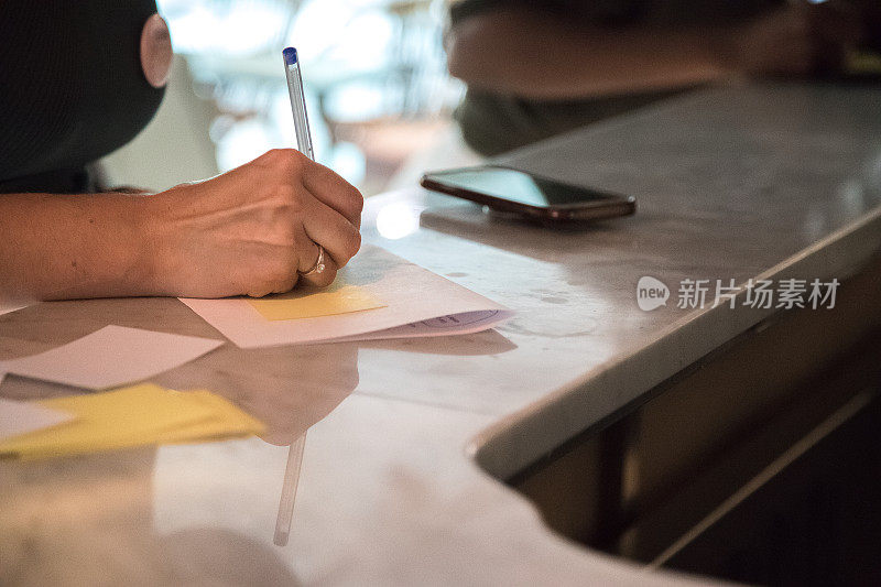 一个女人握着一支笔，在纸上写着一句话。智能手机放在桌子或柜台上。笔迹和新技术的对比。关闭了。
