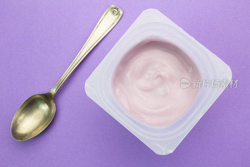 健康的草莓果味酸奶与自然着色在塑料杯孤立的紫色背景与小银匙顶视图