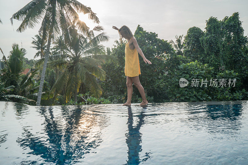 年轻女子走在一个热带天堂目的地的游泳池边缘;女游客在旅游度假池享受度假乐趣的概念