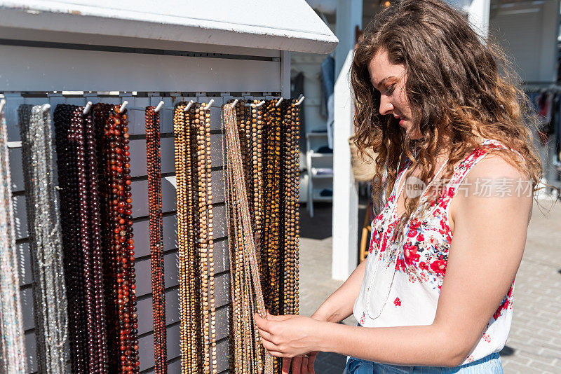 年轻女子购买多彩的石头海滩珠项链触摸珠宝在户外市场商店商店在欧洲，希腊，意大利或地中海城镇村庄在夏天