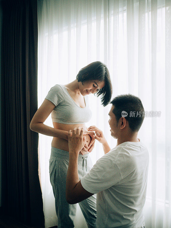 年轻的孕妇和她的丈夫抱着和听婴儿在肚子附近的窗户在家里，怀孕和家庭的概念