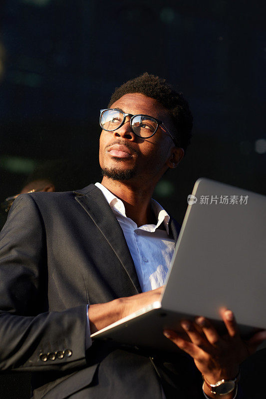 严肃而有目的的年轻黑人商人穿着夹克站在户外，拿着笔记本电脑，想着商业文章
