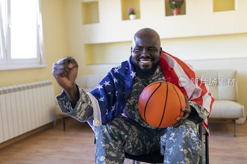 快乐的残疾非裔美国退伍军人坐在轮椅上，他不再现役，他的白人妻子坐在他旁边，在家里的客厅里举着美国国旗为他们的篮球队的胜利欢呼。