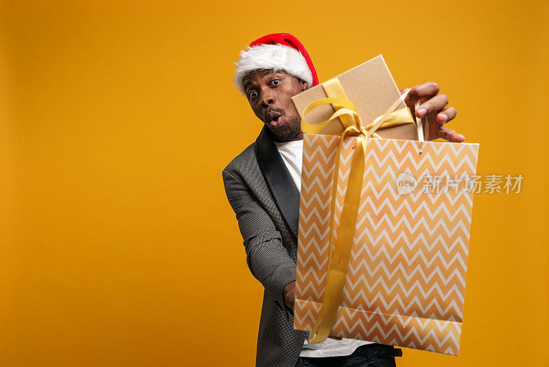 一名戴着圣诞帽的黑人震惊地站在镜头前，拿出一个礼物袋