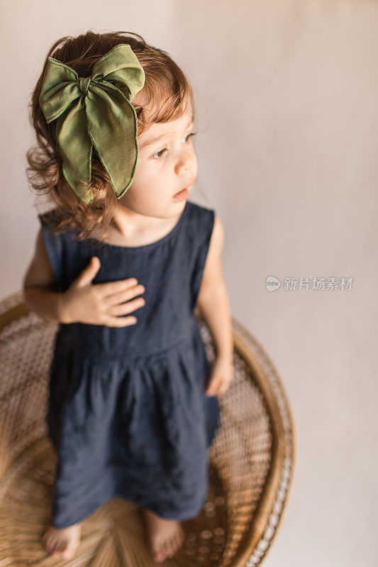 两岁蹒跚学步的小女孩穿着海军蓝亚麻太阳裙和橄榄绿头发蝴蝶结，站在复古藤椅上看窗外