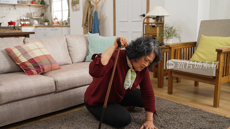 亚洲虚弱的老年妇女试图慢慢起床后，她滑倒在家里客厅的地板上。她用一根棍子支撑自己