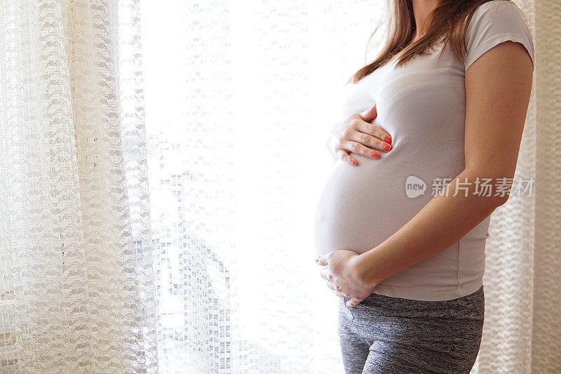 一个孕妇站在窗边，抱着她的肚子，靠得很近