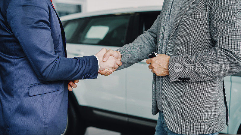 汽车展厅的男工作人员正站在豪华新车旁，将车钥匙交给购车青年，并与他握手。买卖车辆的概念。