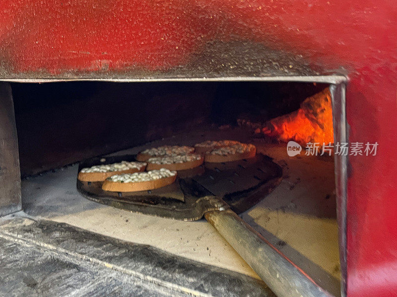 特写形象的迷你披萨被滑入户外，不锈钢，石基础披萨烤箱长柄披萨桨，燃烧的木材，发光的火焰，重点在前景