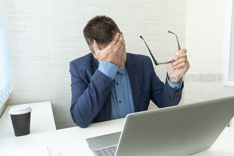 疲惫或筋疲力尽的男人闭着脸，双手坐在办公室的电脑前。商人用手蒙住脸和眼睛。facepalm指