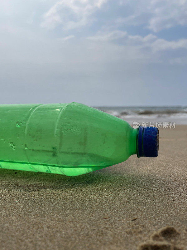 特写图像，空的，绿色塑料饮料瓶与蓝色盖子被冲到沙滩上在退潮水的边缘，破碎的海浪冲刷海洋垃圾和岸上的污染，乱扔的沙子，肮脏的海滩，关注前景，复制空间