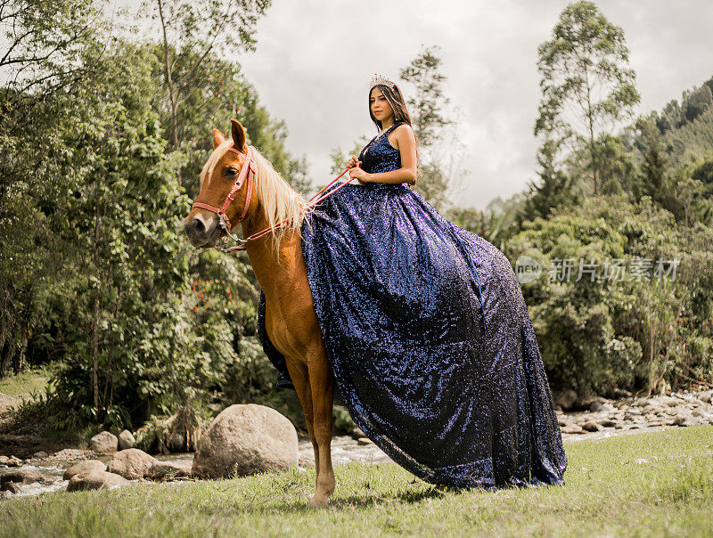 漂亮的年轻女子穿着蓝色的quinceañera连衣裙，头上戴着皇冠，沿着河岸骑马。