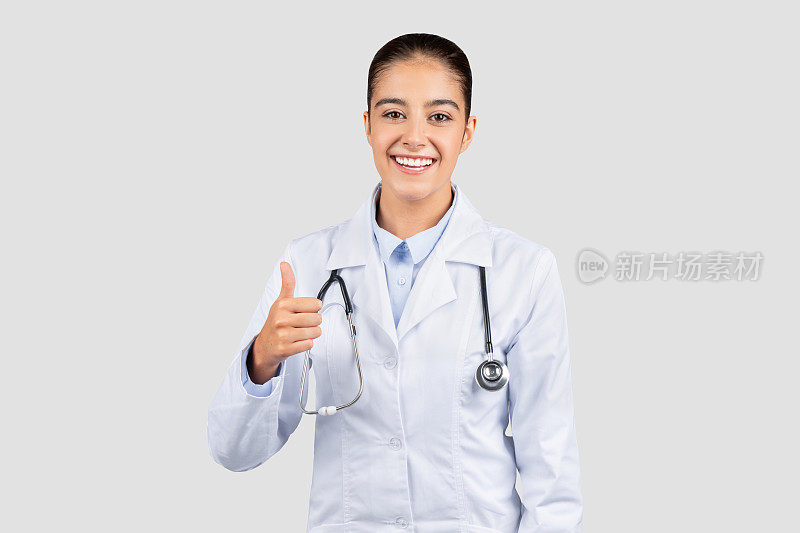 阳性的白人千禧一代医生竖起大拇指，穿着实验室外套，戴着听诊器