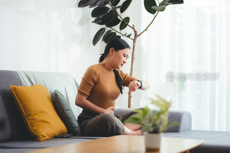 亚洲妇女坐在家里客厅的沙发上感到胃痛。