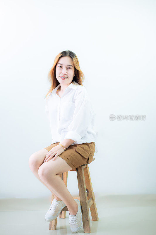 年轻的亚洲女人独自坐在白色背景的椅子上。工作室剪出的美丽短发亚洲女模特的肖像，穿着长袖白衬衫和休闲鞋，盘腿坐着。