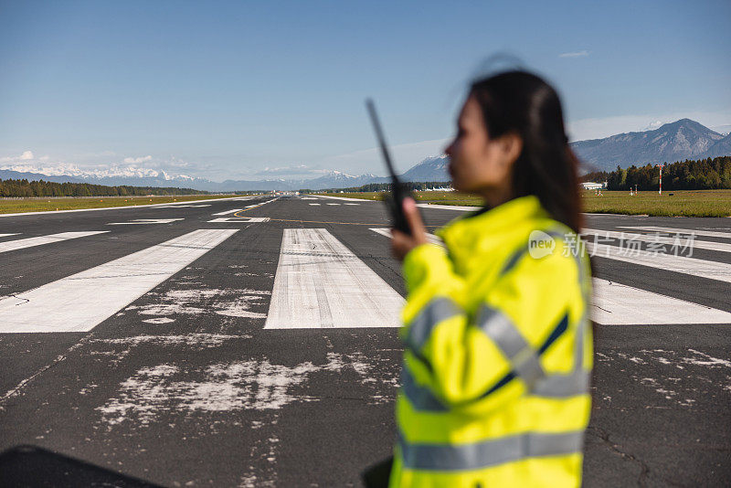 一名成年女机场工作人员的模糊肖像，她穿着黄色安全夹克，在机场跑道中央用对讲机说话