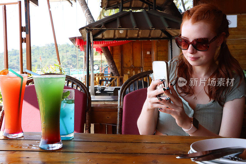 迷人的红发女人用智能手机拍照，彩虹无尾鸡尾酒，蓝色泻湖和龙舌兰日出鸡尾酒，桌子上有塑料吸管，冰块，户外戴太阳镜，户外用餐背景