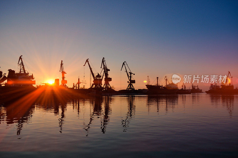 夕阳映衬着繁忙港口的船舶起重机。商业货运码头在黄金时段，太阳反射在水面上。全球贸易、货运、工业天际线、物流基础设施。