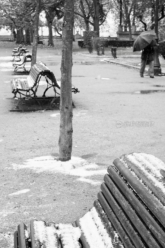 公园长椅上有雪，一个人撑着伞走着。