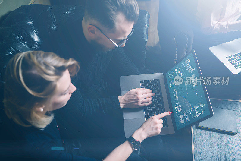 办公室协同工作流程。两个年轻的同事在用电脑。穿着黑色套衫坐在沙发上的女人。图形，在线界面和图表在笔记本电脑屏幕上。水平,视觉效果。