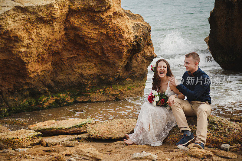 靠近海边的户外沙滩婚礼，时尚幸福的新郎和新娘坐在石头上，有说有笑