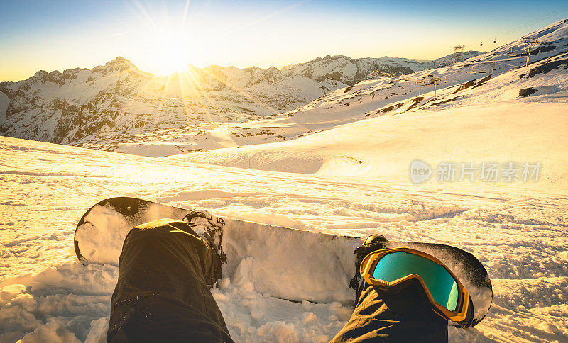 滑雪板坐在放松的时刻在公共滑雪胜地日落-冬季运动概念与人在山顶准备骑下来-腿的观点与温暖的背光滤镜