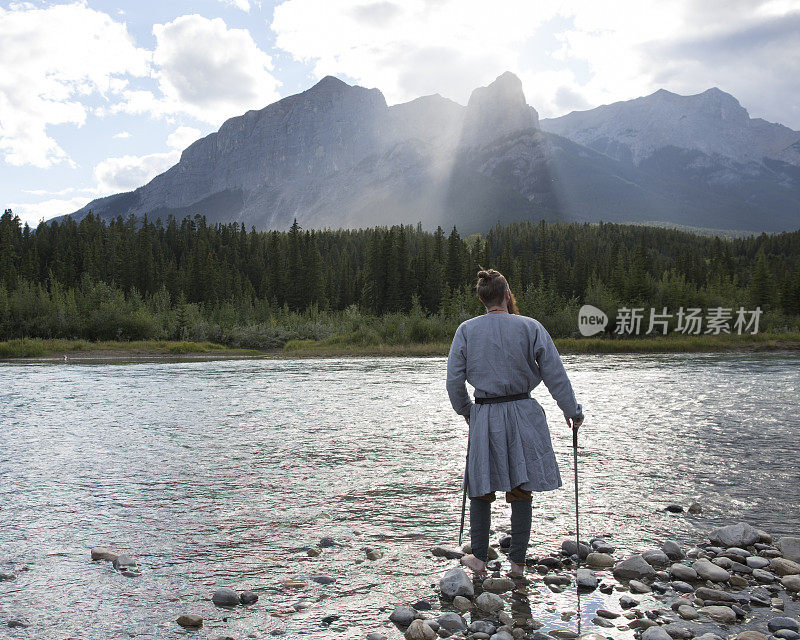 一个穿着诺曼服装的年轻人站在河边，望着群山