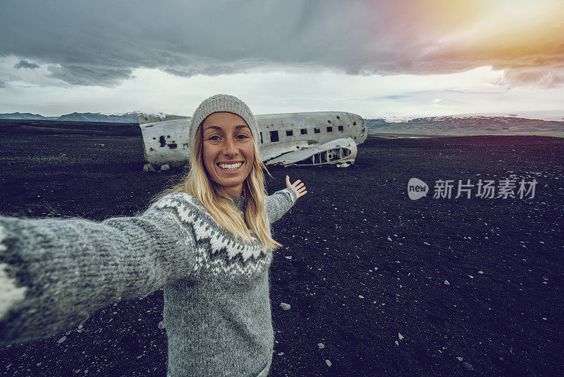 一名年轻女子站在黑沙滩上的飞机残骸旁自拍