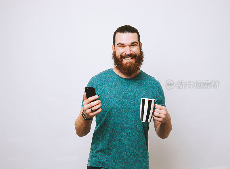 快乐微笑的胡子男人拿着咖啡和智能手机，看着摄像头
