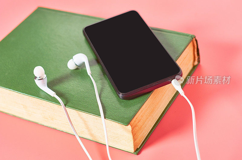 带耳机的智能手机或平板电脑，粉红色背景的绿色旧书。有声读物的概念。在线教育电子学习。现代科技