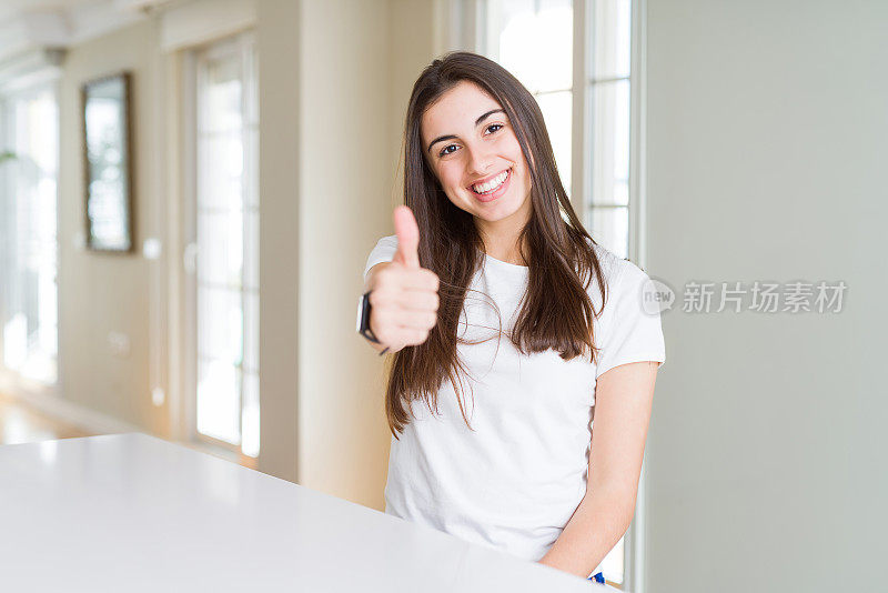 美丽的年轻女子穿着休闲白色t恤做快乐的拇指手势与手。赞许的表情看着镜头，表示成功。